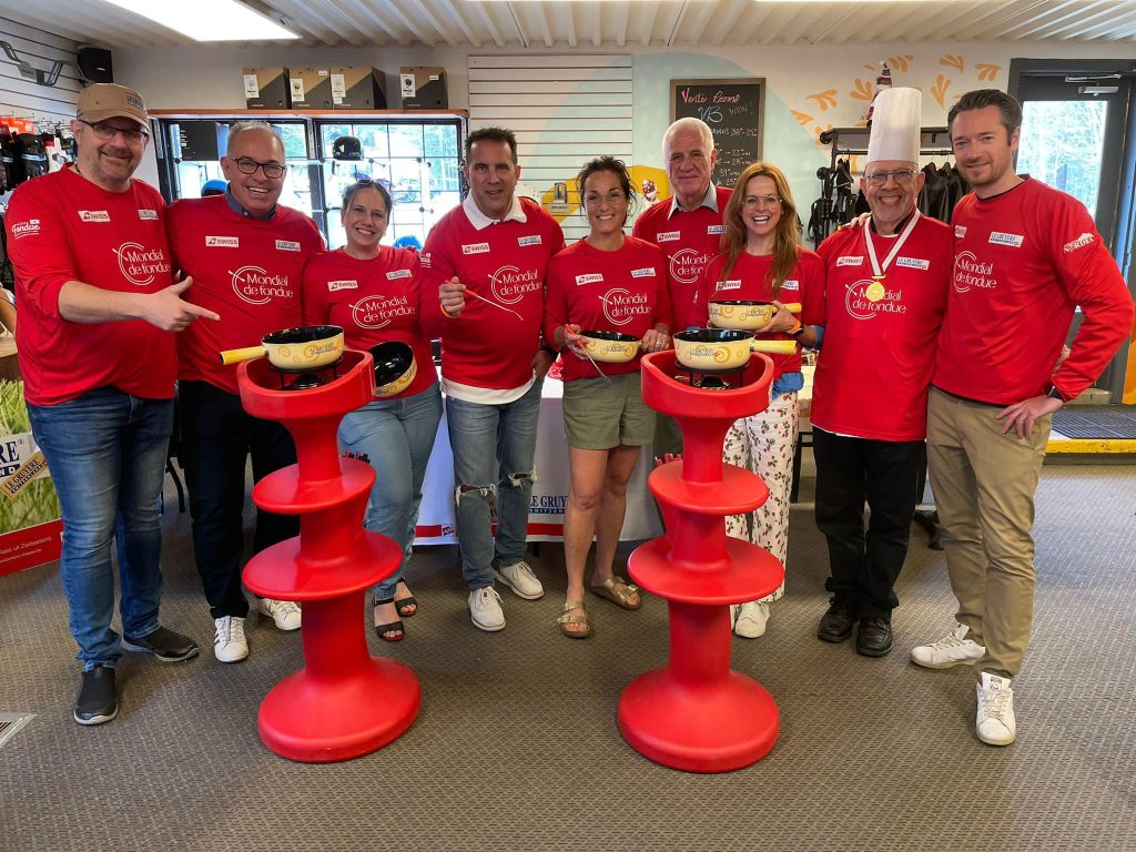 9 personnes souriantes portant un chandail rouge se tiennent debut derrière des plats à fondue au fromage